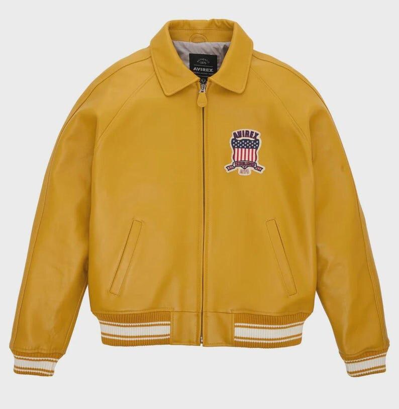 Puede incluir: Chaqueta de cuero amarillo mostaza con cremallera frontal, cuello y pu&ntilde;os y dobladillo acanalados. Cuenta con un parche en el pecho con un dise&ntilde;o de escudo y la palabra "AVIREX". La chaqueta tiene dos bolsillos delanteros.