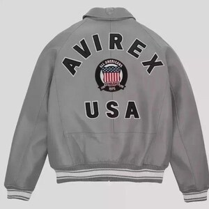 Puede incluir: Chaqueta bomber gris con la palabra "AVIREX" arqueada en la parte superior de la espalda. Debajo, un parche circular con las palabras "ALL AMERICANS" y "ESTABLISHED 1975" que rodea un dise&ntilde;o de la bandera estadounidense. La palabra "USA" est&aacute; debajo.