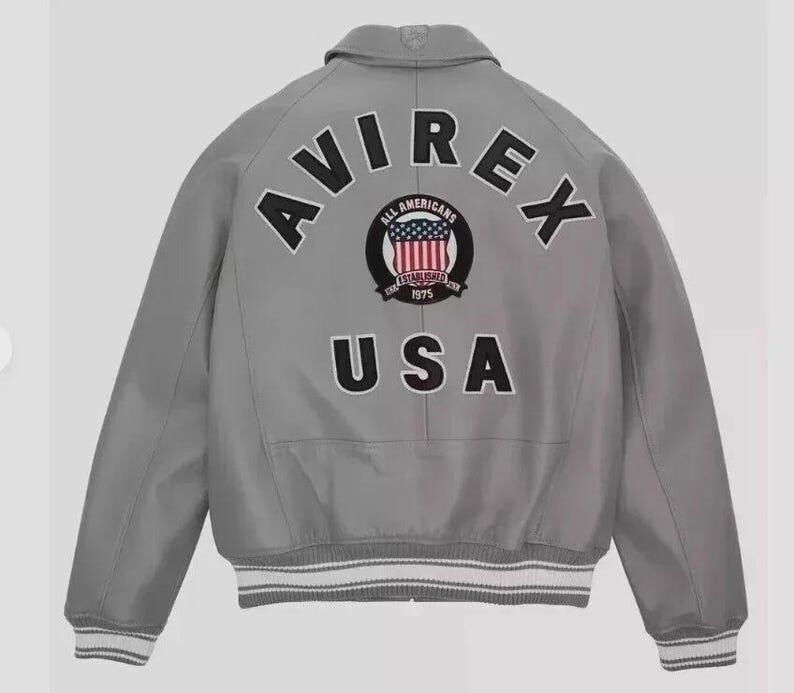 Puede incluir: Chaqueta bomber gris con la palabra "AVIREX" arqueada en la parte superior de la espalda. Debajo, un parche circular con las palabras "ALL AMERICANS" y "ESTABLISHED 1975" que rodea un dise&ntilde;o de la bandera estadounidense. La palabra "USA" est&aacute; debajo.
