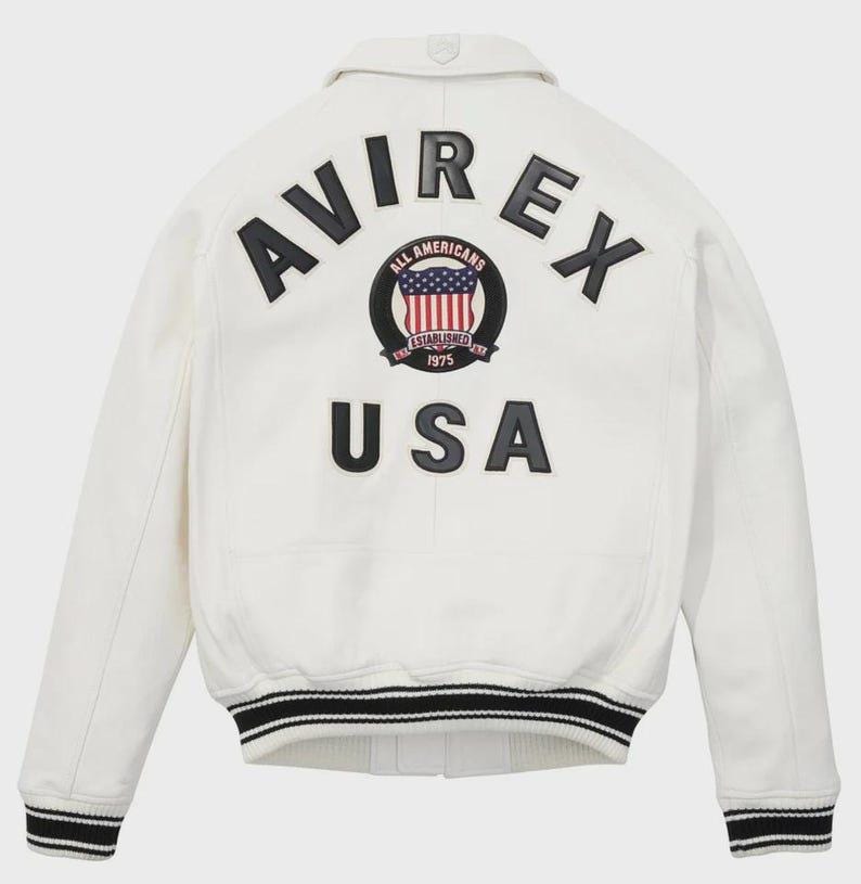 Puede incluir: Chaqueta bomber blanca con ribetes a rayas negras y blancas. La parte trasera presenta la palabra "AVIREX" sobre un parche circular con un dise&ntilde;o de bandera estadounidense y las palabras "ALL AMERICANS ESTABLISHED 1975". Debajo, la palabra "USA".