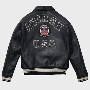 Puede incluir: Chaqueta de cuero negra con la palabra "AVIREX" en letras blancas en la espalda. Debajo, un parche con la bandera americana y las palabras "ALL AMERICANS ESTABLISHED 1975". La palabra "USA" est&aacute; debajo. La chaqueta tiene cuello y pu&ntilde;os a rayas.