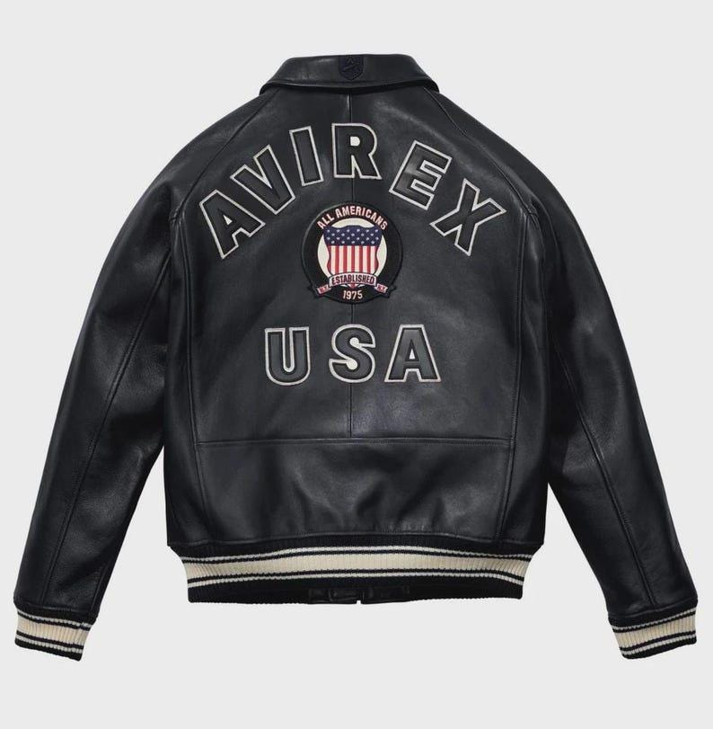 Puede incluir: Chaqueta de cuero negra con la palabra "AVIREX" en letras blancas en la espalda. Debajo, un parche con la bandera americana y las palabras "ALL AMERICANS ESTABLISHED 1975". La palabra "USA" est&aacute; debajo. La chaqueta tiene cuello y pu&ntilde;os a rayas.