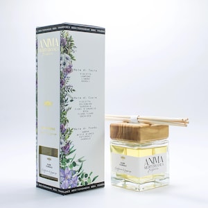 Może przedstawiać: Przezroczysty szklany dyfuzor z drewnianą nasadką i patyczkami, obok wysokiego, prostokątnego pudełka. Pudełko zawiera kwiatowe wzory i tekst, w tym "ANIMA Mediterranea" i nuty zapachowe. Butelka zawiera żółty płyn.