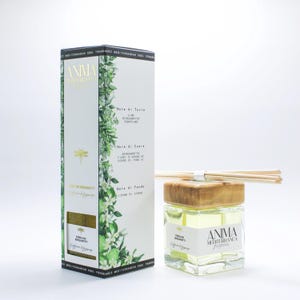 Może przedstawiać: Kwadratowa szklana butelka z drewnianą nakrętką i patyczkami, obok białego pudełka z zielonymi ilustracjami botanicznymi. Butelka zawiera żółty płyn i ma napis "ANIMA Mediterranea". Pudełko zawiera tekst w języku włoskim.