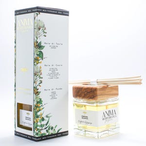 Może przedstawiać: Dyfuzor zapachowy Anima Mediterranea z drewnianym blatem i patyczkami. Przezroczysta szklana butelka zawiera żółty płyn. Obok butelki znajduje się pasujące pudełko z kwiatowymi i botanicznymi ilustracjami.