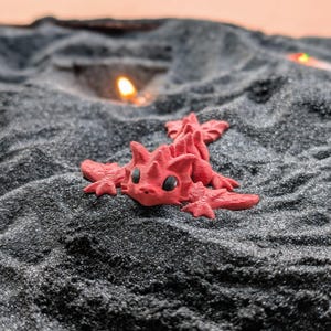 Pode incluir: Uma pequena figura de dragão vermelho com olhos pretos e escamas detalhadas repousa sobre uma cama de areia cinza escuro. O dragão está deitado, com as asas e a cauda abertas. O fundo está desfocado, com um brilho quente e alaranjado.