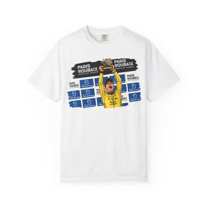 Puede incluir: Camiseta blanca con un gráfico de un ciclista con un maillot amarillo sosteniendo un trofeo, con el texto "PARIS ROUBAIX HAUTS-DE-FRANCE". El diseño incluye detalles en azul y negro, y la camiseta tiene cuello redondo.