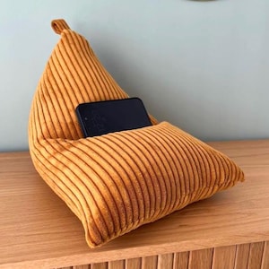 Op de afbeelding: Een mosterdkleurige corduroy telefoonstandaard met een vak om een telefoon in te plaatsen. De standaard is driehoekig van vorm met een afgeronde bovenkant en een rechthoekige basis. De telefoon is zwart.