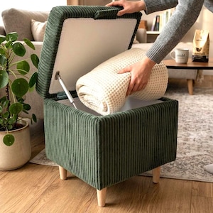 Op de afbeelding: Een vierkante opberghocker bekleed met donkergroene corduroy stof met vier lichtgekleurde houten poten. Het deksel is scharnierend en geopend, waardoor een holle binnenkant zichtbaar is met een gevouwen crèmekleurige wafelstructuurdeken.