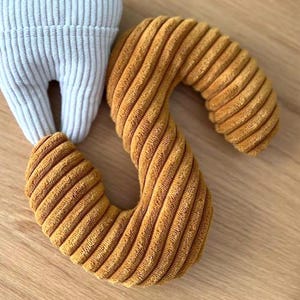 Può includere: Un giocattolo di peluche color bianco sporco a forma di dente con una consistenza a coste, posizionato accanto a un giocattolo di peluche color giallo senape a forma di S con una struttura in tessuto a coste simile. Entrambi sono oggetti morbidi.