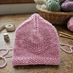 May include: A hand-knitted pink baby hat with a heart design. The hat is made of soft yarn and sits on a wooden surface. A basket of yarn balls in various colors and knitting needles are in the background. The text "Knit with Love" is visible.