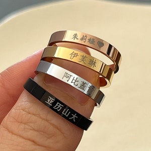 May include: A stack of five rings in various metallic finishes: rose gold, gold, silver, and black. Each ring has engraved text in a foreign language. The rings are stacked on a finger against a blurred background.