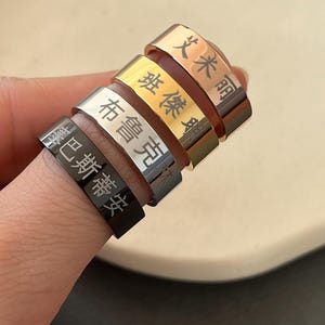 May include: A stack of four rings in varying metallic finishes: black, silver, gold, and rose gold. Each ring features engraved characters. The rings are stacked on a finger against a neutral background.