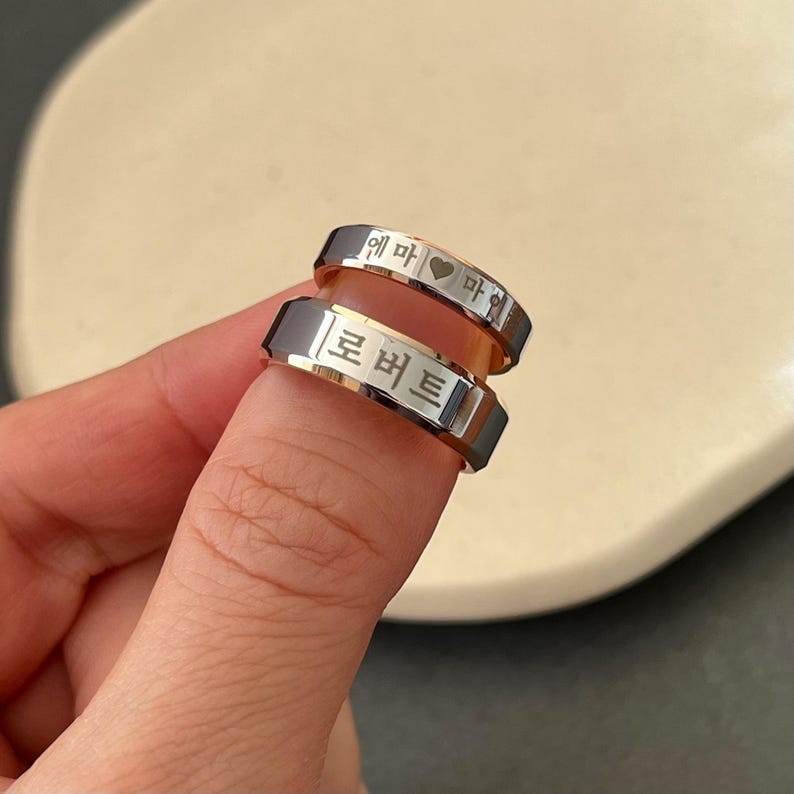 May include: Two silver rings with engraved Korean text. The top ring has a heart symbol and the text "에마 &hearts; 마이". The bottom ring has the text "프버트".