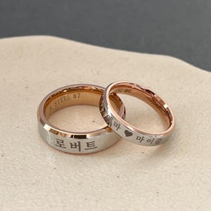 May include: Two stainless steel wedding bands with rose gold accents. The band on the left has the inscription "S. STEEL #7" and the Korean word "로버트". The band on the right has the inscription "S. STEEL #6" and the Korean words "혜마 &hearts; 마이클".