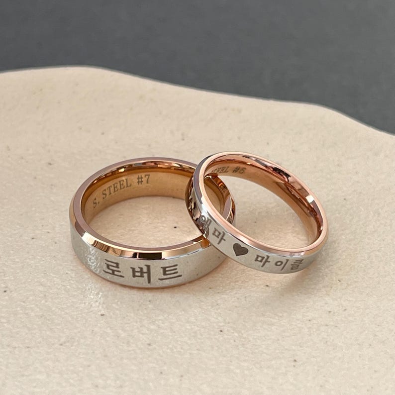 May include: Two stainless steel wedding bands with rose gold accents. The band on the left has the inscription "S. STEEL #7" and the Korean word "로버트". The band on the right has the inscription "S. STEEL #6" and the Korean words "혜마 &hearts; 마이클".