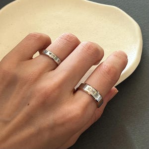 May include: Two silver rings with Korean text engraved on them. The rings are worn on the fingers of a person's hand.