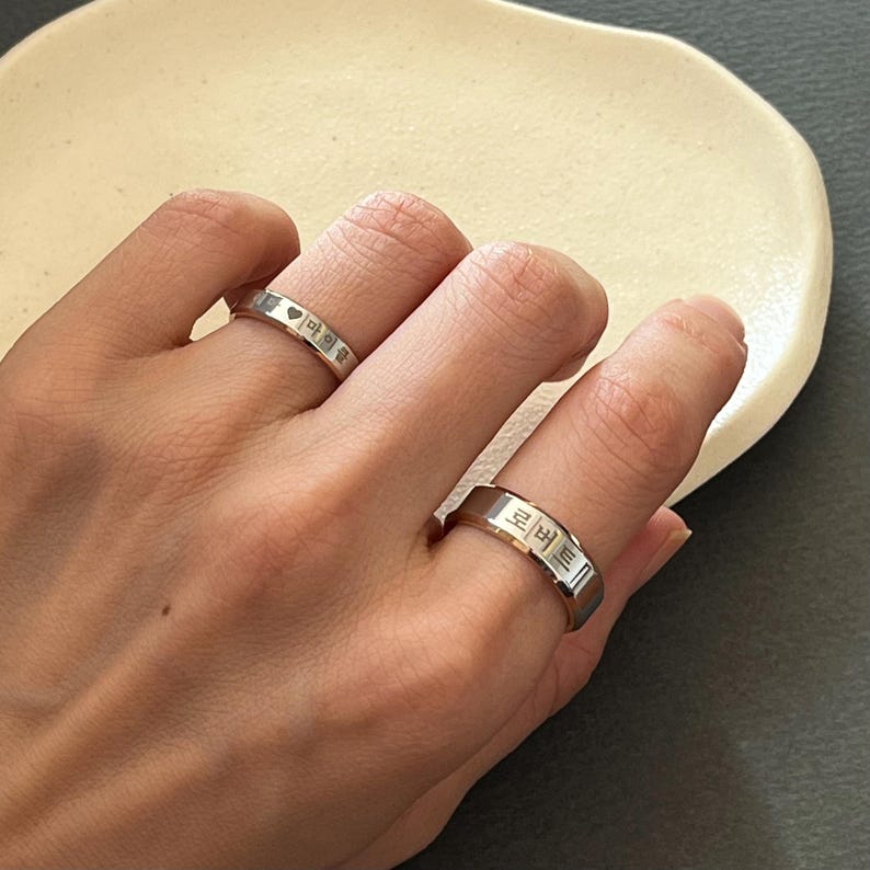 May include: Two silver rings with Korean text engraved on them. The rings are worn on the fingers of a person's hand.