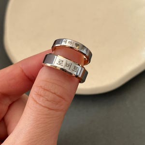May include: Two silver rings with engraved Korean text. The top ring has the text "에마" and a heart symbol. The bottom ring has the text "로버트".