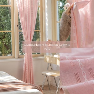 May include: Pink light filtering curtain with a textured design, hanging in front of a window. The curtain has a delicate, semi-sheer appearance, allowing soft light to filter through. The text on the curtain reads "CAMILLE LIGHT FILTERING CURTAIN PINK".