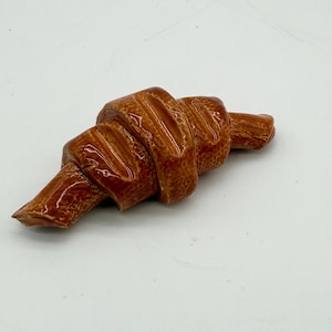 May include: A ceramic croissant sculpture in shades of brown. The pastry is detailed with ridges and a glossy finish, suggesting a baked texture. The sculpture is a decorative item, likely for display.