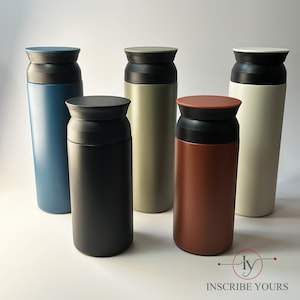 May include: Five insulated travel mugs in various colors: blue, black, olive green, rust, and cream. Each mug has a black lid and a matching colored base. The mugs are cylindrical and appear to be made of metal. The logo "INSCRIBE YOURS" is in the bottom right corner.