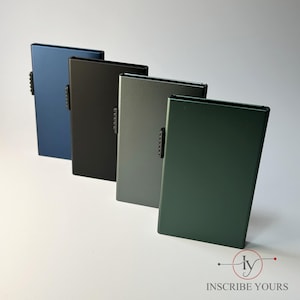 Puede incluir: Cinco tarjeteros rectangulares en tonos azul, negro, gris y verde. Cada uno tiene un pequeño cierre metálico texturizado. Los tarjeteros están dispuestos sobre una superficie blanca. La marca "INSCRIBE YOURS" está en la parte inferior.