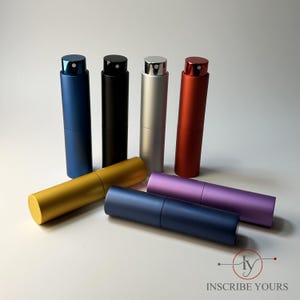 May include: A collection of seven cylindrical metal perfume atomizer spray bottles in various matte colors including blue, black, silver, red, gold, purple, and dark blue, with the text INSCRIBE YOURS and a logo in the bottom right corner.