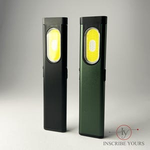May include: Two rectangular LED work lights. One is black, the other is dark green. Each has a black top and a bright yellow, oval-shaped light. The lights are standing upright against a neutral background. The text "INSCRIBE YOURS" is in the bottom right corner.