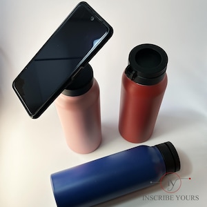 Könnte beinhalten: Drei zylindrische Wasserflaschen mit schwarzen Deckeln in mattem Rosa, Dunkelrot und Marineblau. Ein schwarzes Smartphone ist schräg auf der rosa Flasche balanciert. Der Text "INSCRIBE YOURS" und ein kreisförmiges Logo sind unten rechts zu sehen.