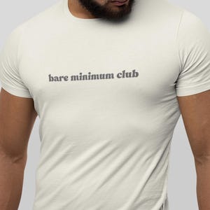 May include: Cream-colored t-shirt with the text "bare minimum club" printed in gray. The shirt has short sleeves and a crew neck. The fabric appears to be soft and comfortable.