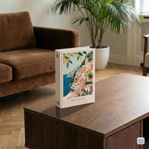 May include: A hardback book with a colourful illustration of a coastal Italian town. The cover features a vibrant scene with buildings, the sea, and lemon branches. The book is on a dark brown wooden table.