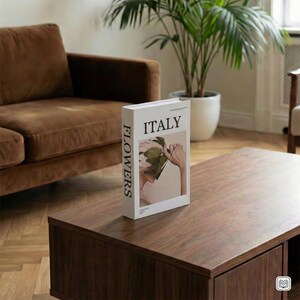 Puede incluir: Un libro blanco titulado "ITALY" con la palabra "FLOWERS" impresa en el lomo, apoyado sobre una mesa de centro de madera oscura. La portada del libro presenta el rostro de una mujer parcialmente oculto por hojas. Un sofá marrón y una planta en maceta están en el fondo.