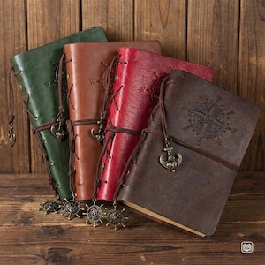 Op de afbeelding: Een verzameling lederen notitieboeken in groen, bruin en rood. Elk notitieboek heeft een decoratieve bedel en een leren sluiting. De notitieboeken hebben een nautisch thema, met kompas- en ankerbedels.