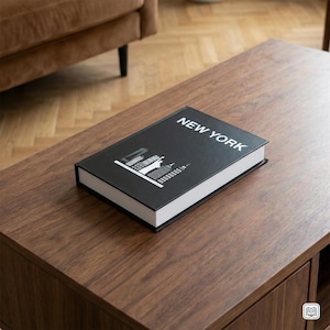 Op de afbeelding: Een zwart boek met de woorden "NEW YORK" in witte letters. De omslag toont een witte en grijze skyline-illustratie. Het boek ligt op een donkerbruine houten salontafel.