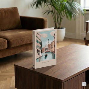 Può includere: Un libro con copertina rigida con un'illustrazione di Venezia. La copertina presenta una scena del canale con edifici, un ponte e una gondola. Il libro è su un tavolo di legno marrone scuro, con la parola "Venezia" stampata sulla copertina.
