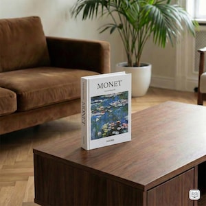 Könnte beinhalten: Ein Buch mit dem Titel "MONET" liegt auf einem dunklen Holztisch. Das Cover des Buches zeigt ein Seerosen-Gemälde. Das Buch ist weiß mit schwarzem Text. Ein braunes Sofa und eine Topfpflanze sind im Hintergrund zu sehen.
