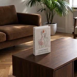 May include: A white decorative book titled GALERIE BOHÈME with a black line art illustration of a person's back and head on the cover. The cover features a small orange circular accent and a soft pink shape, with black text printed below the illustration.