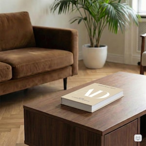 Könnte beinhalten: Ein dunkelbrauner Couchtisch mit einem Buch mit dem Titel "AD A Century of Style". Das Buch hat einen beigefarbenen Einband und weiße Schrift. Ein braunes Sofa und eine Topfpflanze bilden den Hintergrund.