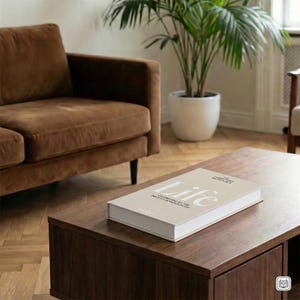 Puede incluir: Una mesa de centro de madera marrón oscuro con un libro titulado "Life" encima. El libro tiene una cubierta beige con texto blanco. Un sofá marrón y una planta en maceta están en el fondo. La mesa tiene un estante inferior.