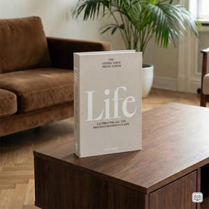 May include: A beige hardback photo album titled "Life" with the text "Celebrating All The Precious Moments In Life." The album is on a dark brown wooden coffee table. A brown sofa and a potted plant are in the background.