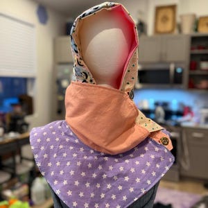 May include: A hooded scarf with a celestial pattern, peach-colored lining, and a lavender bib adorned with white stars. The scarf is secured with two decorative buttons, offering a blend of style and warmth.