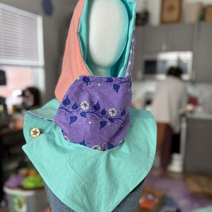 May include: A handmade hood with a variety of fabrics. The hood features a light pink exterior, a teal interior, and a purple neck piece with a floral pattern. A wooden star-shaped button secures the neck piece.