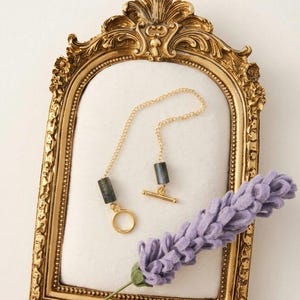 May include: A gold-toned necklace with a bar and ring clasp, featuring dark gray rectangular beads. The necklace is displayed inside a gold ornate frame, with a purple felt flower stem in the foreground.