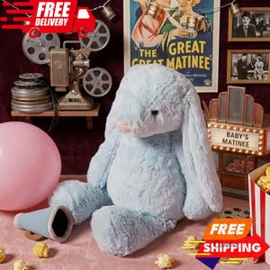 May include: A large, light blue plush bunny with long ears and a pink nose. The bunny is sitting on a pink surface, with a blue megaphone and a pink balloon nearby. The background includes a vintage movie poster and a popcorn box.