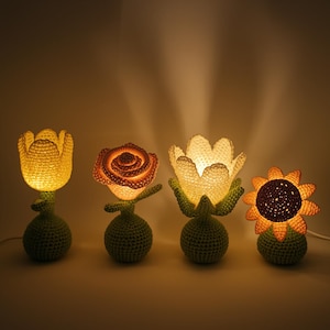 May include: Four crochet flower lamps, each with a unique design: a tulip, a rose, a lily, and a sunflower. The lamps are illuminated, producing a warm glow. The bases and stems are green, and the flowers are various shades of yellow and orange.