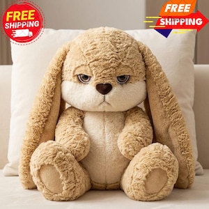 May include: A plush, light brown bunny with long, floppy ears and a slightly grumpy expression. The stuffed animal has a heart-shaped nose and is sitting on a white pillow. The bunny's fur is soft and textured, with a lighter cream color on its face and belly.