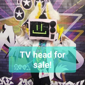 May include: A costume headpiece shaped like a light pink television set with a black screen featuring a bright green smiley face. The headpiece includes tall white and yellow faux fur ears, yellow lightning bolt side accents, and two black knobs.
