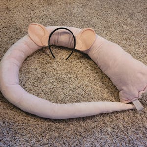 May include: A light pink plush costume set consisting of a headband with two rounded ears and a long, stuffed, curved tail. The tail features two white elastic loops at one end for attachment, and the entire set is made of a soft, fuzzy fabric.