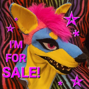 May include: A colorful animal head mask featuring bright blue and yellow fabric, a vibrant pink faux fur mohawk, and sharp teeth. The mask has large pointed ears, a black nose, and pink eyes with mesh detailing. The text I'M FOR SALE! is displayed.